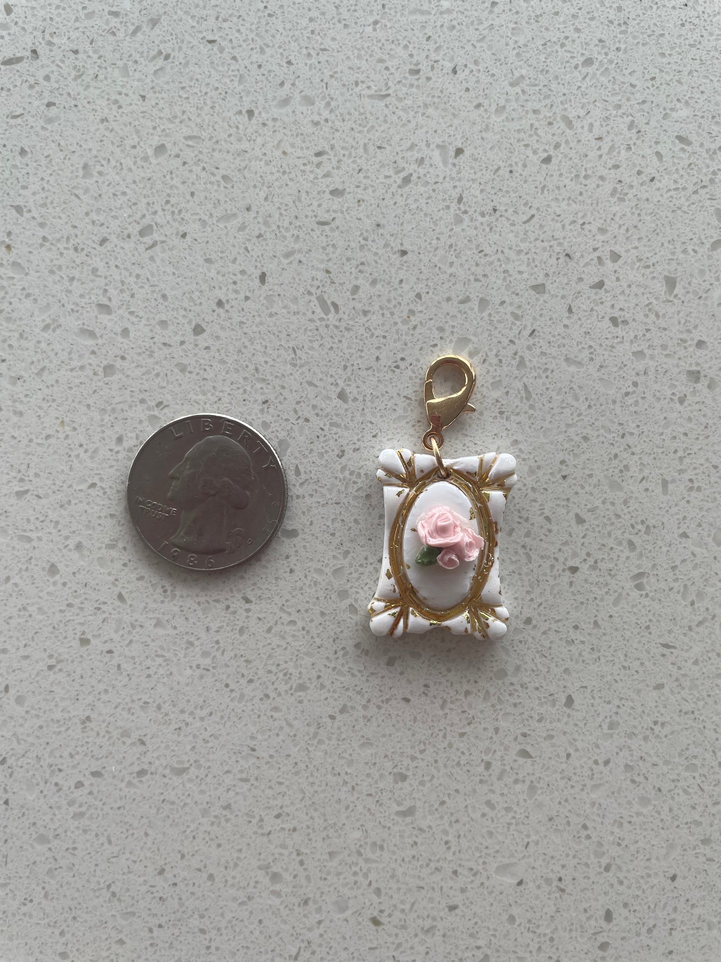 Miniature decorative charm with a pink flower and gold frame on a gray surface, next to a coin for scale.