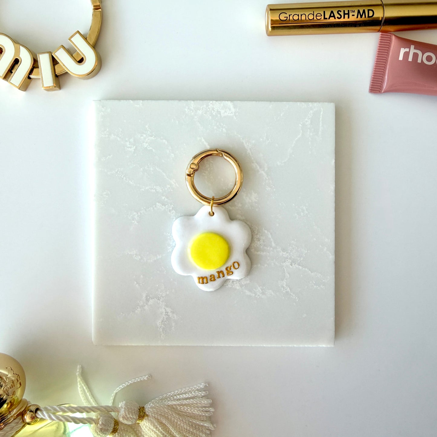 A pet tag in the shape of a white daisy flower with a bright yellow center lays on a marble surface. It has the word "mango" in lowercase gold lettering across the lower right corner and is attach to a gold ring clasp. It is staged on a surface next to various cosmetics and charms.