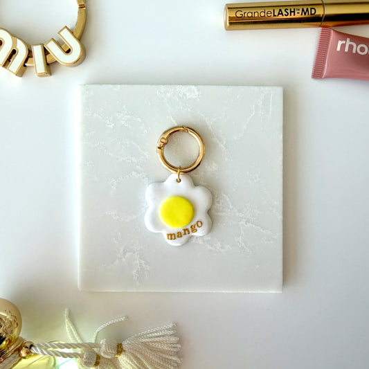 A pet tag in the shape of a white daisy flower with a bright yellow center lays on a marble surface. It has the word "mango" in lowercase gold lettering across the lower right corner and is attach to a gold ring clasp. It is staged on a surface next to various cosmetics and charms.