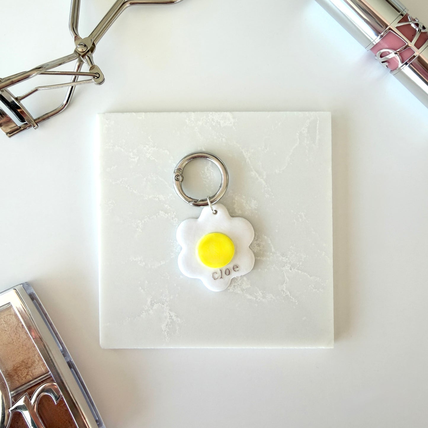 A pet tag in the shape of a white daisy flower with a bright yellow center lays on a marble surface. It has the word "cloe" in lowercase silver lettering across the lower right corner and is attach to a silver ring clasp. It is staged on a surface next to various cosmetics.