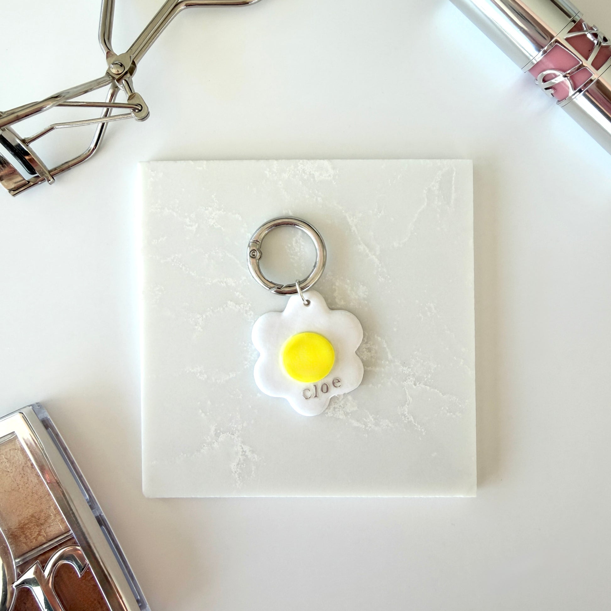 A pet tag in the shape of a white daisy flower with a bright yellow center lays on a marble surface. It has the word "cloe" in lowercase silver lettering across the lower right corner and is attach to a silver ring clasp. It is staged on a surface next to various cosmetics.