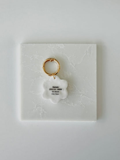 A pet tag in the shape of a white daisy flower flipped over so the back face is visible. It has the words " Example (555) 555 - XXXX 555 Street City, State" in small black print.