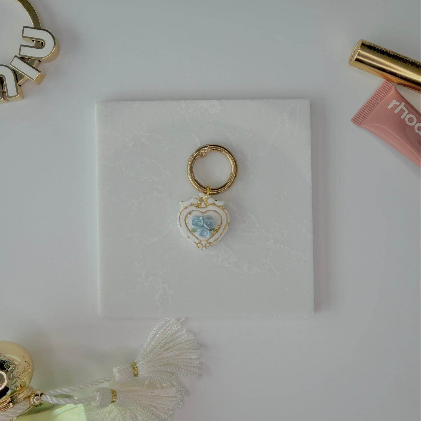 Heart-shaped pet ID tag with blue roses on a gold ring sitting on a marble square staged next to a rhode peptide lip treatment, gold miu miu bag charm, gold lash serum, and a Parfum de Marly bottle.