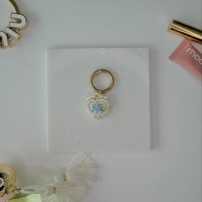 Heart-shaped pet ID tag with blue roses on a gold ring sitting on a marble square staged next to a rhode peptide lip treatment, gold miu miu bag charm, gold lash serum, and a Parfum de Marly bottle.