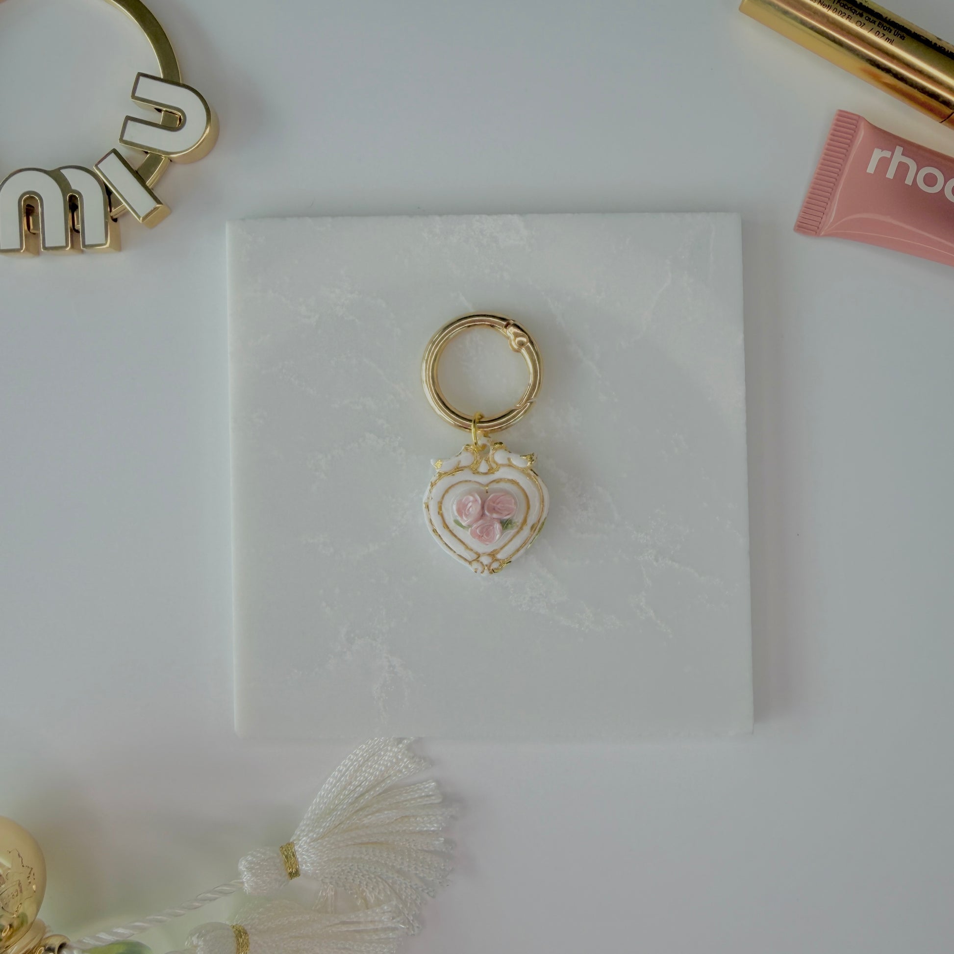 Heart-shaped pet ID tag with pink roses on a gold ring sitting on a marble square staged next to a rhode peptide lip treatment, gold miu miu bag charm, gold lash serum, and a Parfum de Marly bottle.