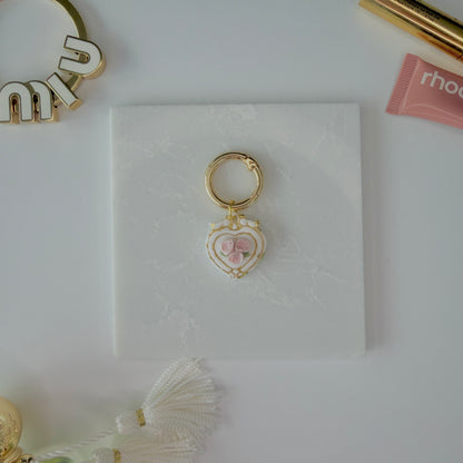 Heart-shaped pet ID tag with pink roses on a gold ring sitting on a marble square staged next to a rhode peptide lip treatment, gold miu miu bag charm, gold lash serum, and a Parfum de Marly bottle.