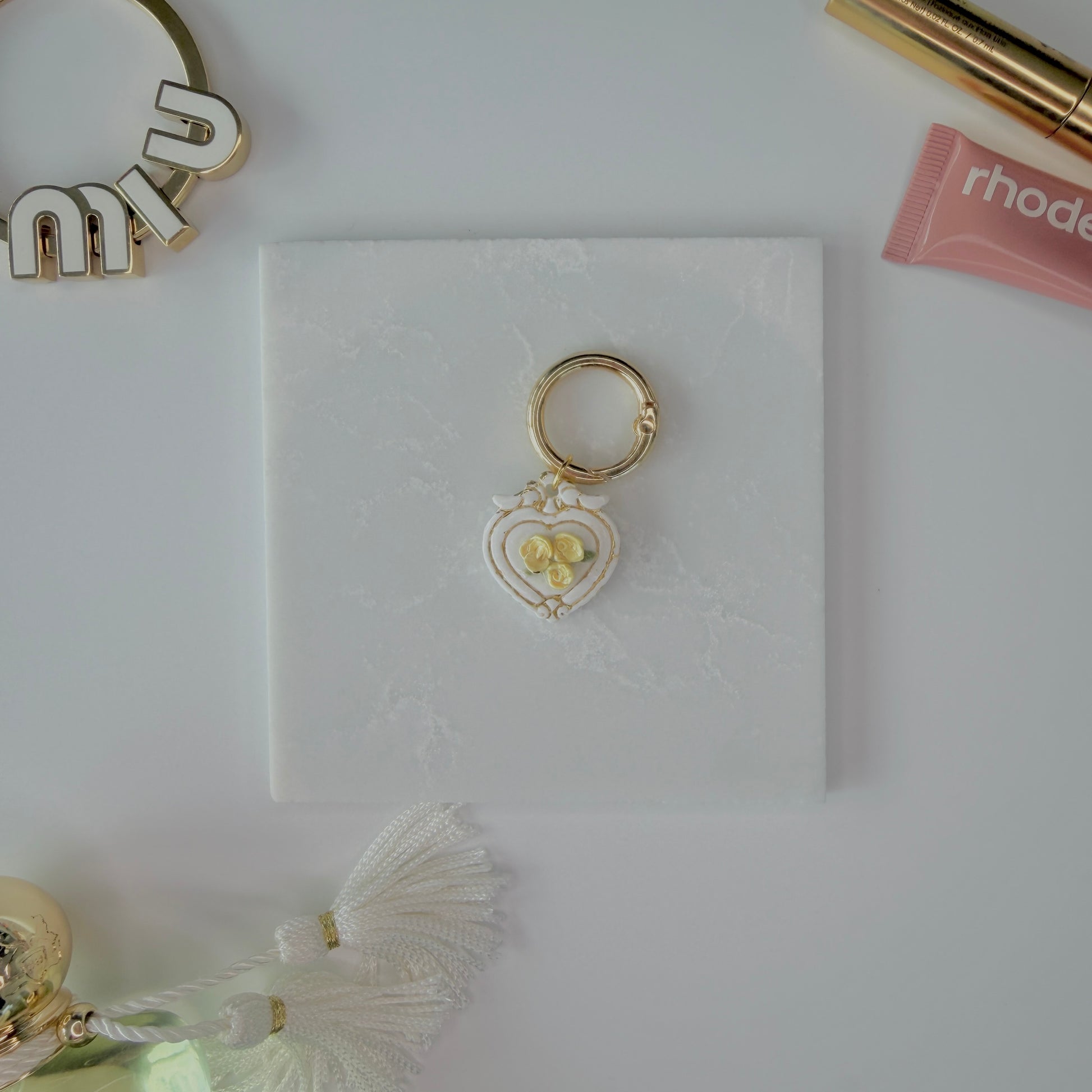 Heart-shaped pet ID tag with yellow roses on a gold ring sitting on a marble square staged next to a rhode peptide lip treatment, gold miu miu bag charm, gold lash serum, and a Parfum de Marly bottle.