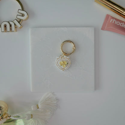 Heart-shaped pet ID tag with yellow roses on a gold ring sitting on a marble square staged next to a rhode peptide lip treatment, gold miu miu bag charm, gold lash serum, and a Parfum de Marly bottle.