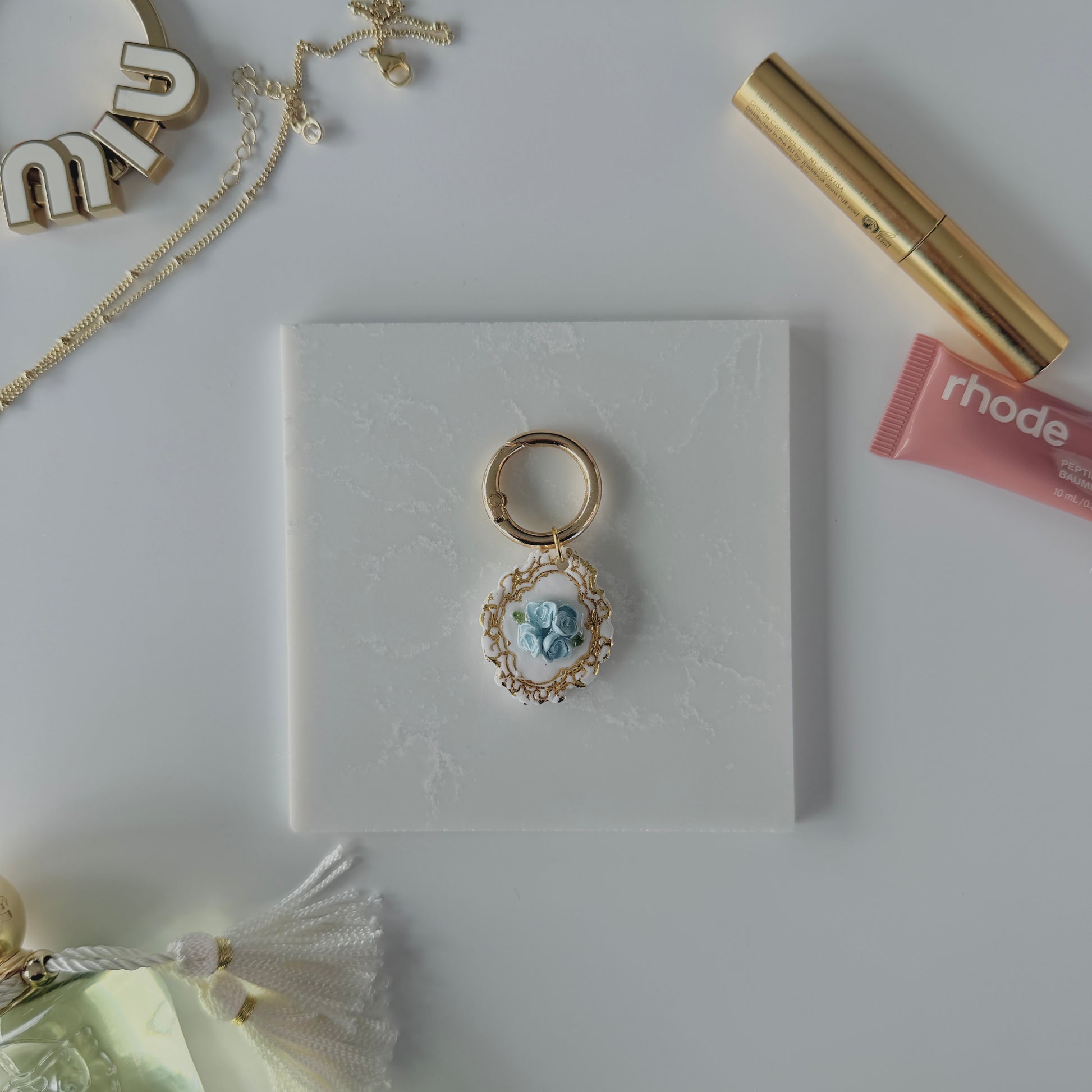 A product image of a white pet ID Tag with four blue roses and gold engraving attached to a gold ring sits on a piece of marble. Staged around it are a miu miu bag charm, gold necklace, rhode lip gloss Parfum de Marly perfume bottle, and a gold lash serum all on a white background.
