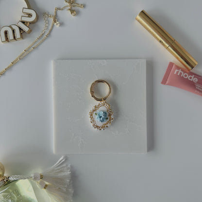 A product image of a white pet ID Tag with four blue roses and gold engraving attached to a gold ring sits on a piece of marble. Staged around it are a miu miu bag charm, gold necklace, rhode lip gloss Parfum de Marly perfume bottle, and a gold lash serum all on a white background.