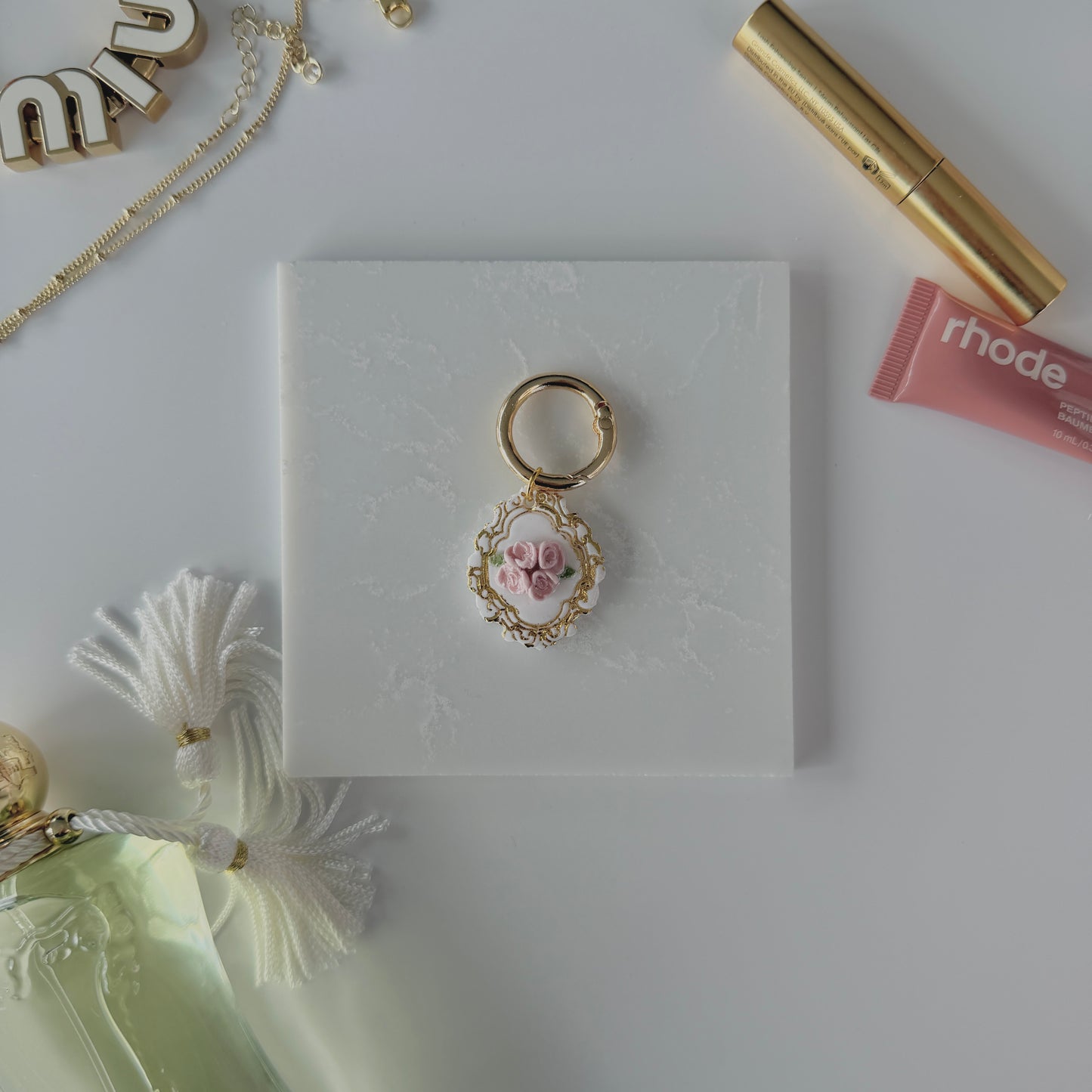 A product image of a white pet ID Tag with four pink roses and gold engraving attached to a gold ring sits on a piece of marble. Staged around it are a miu miu bag charm, gold necklace, rhode lip gloss,  Parfum de Marly perfume bottle, and a gold lash serum all on a white background.