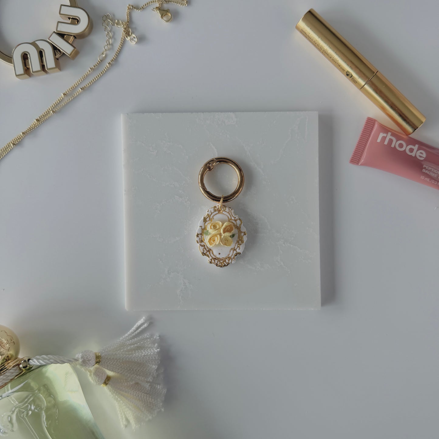 A product image of a white pet ID Tag with four yellow roses and gold engraving attached to a gold ring sits on a piece of marble. Staged around it are a miu miu bag charm, gold necklace, rhode lip gloss  Parfum de Marly perfume bottle, and a gold lash serum all on a white background.