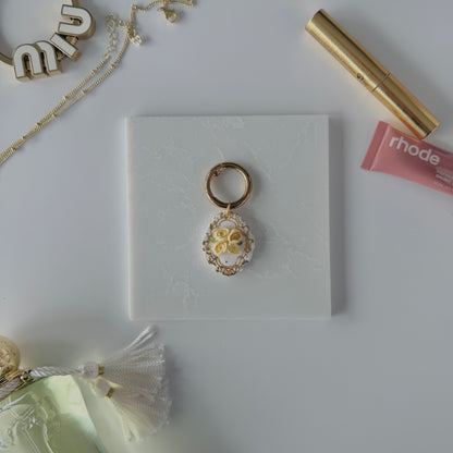 A product image of a white pet ID Tag with four yellow roses and gold engraving attached to a gold ring sits on a piece of marble. Staged around it are a miu miu bag charm, gold necklace, rhode lip gloss  Parfum de Marly perfume bottle, and a gold lash serum all on a white background.