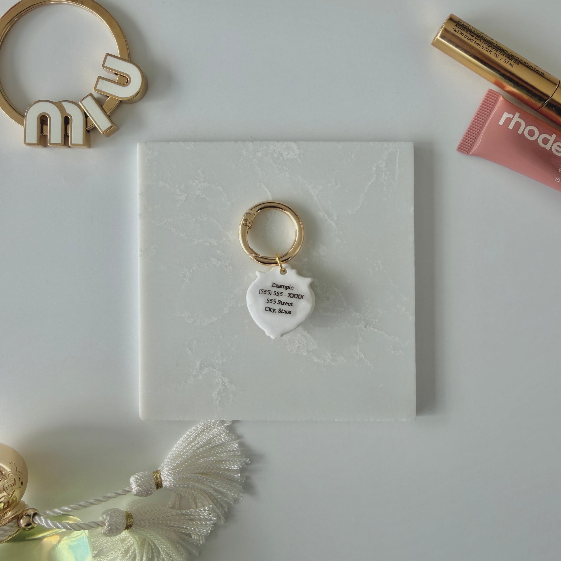 Product image. Back face of a heart-shaped pet ID tag on a gold ring sitting on a marble square. The label reads: Example (555) 555- XXXX 555 Street City, State. It is staged next to cosmetics, bag charms, and a perfume bottle.