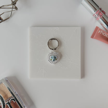 Heart-shaped pet ID tag with blue roses on a silver ring sitting on a marble square staged next to a rhode peptide lip treatment, YSL lipstick, silver E.L.F eyelash curler, and a Dior highlighting kit.
