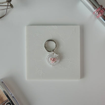 Heart-shaped pet ID tag with pink roses on a silver ring sitting on a marble square staged next to a YSL lipstick, silver E.L.F eyelash curler, and a Dior highlighting palette.