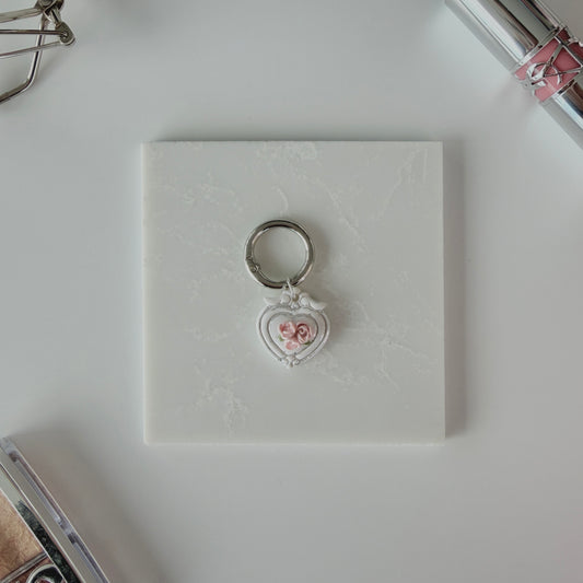 Heart-shaped pet ID tag with pink roses on a silver ring sitting on a marble square staged next to a YSL lipstick, silver E.L.F eyelash curler, and a Dior highlighting palette.