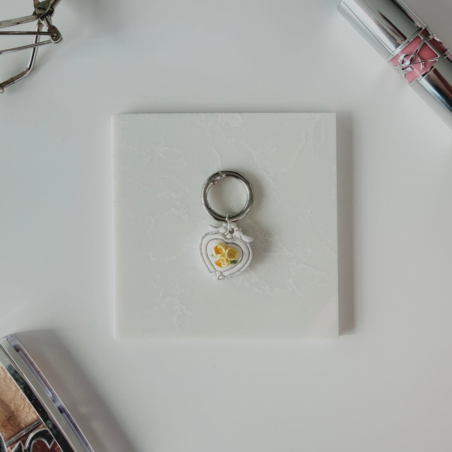 Heart-shaped pet ID tag with yellow roses on a silver ring sitting on a marble square staged next to a YSL lipstick, silver E.L.F eyelash curler, and a Dior highlighting palette.