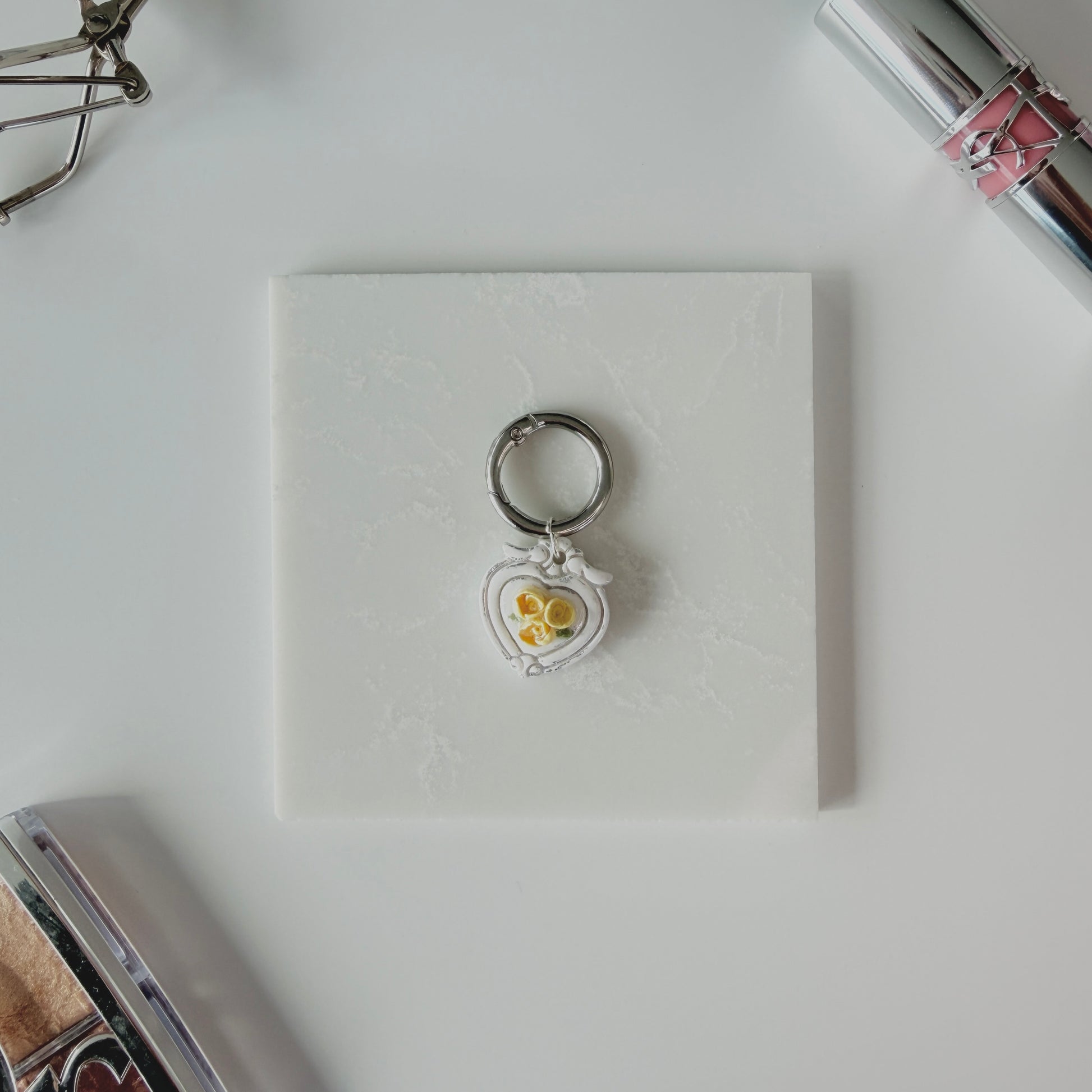 Heart-shaped pet ID tag with yellow roses on a silver ring sitting on a marble square staged next to a YSL lipstick, silver E.L.F eyelash curler, and a Dior highlighting palette.