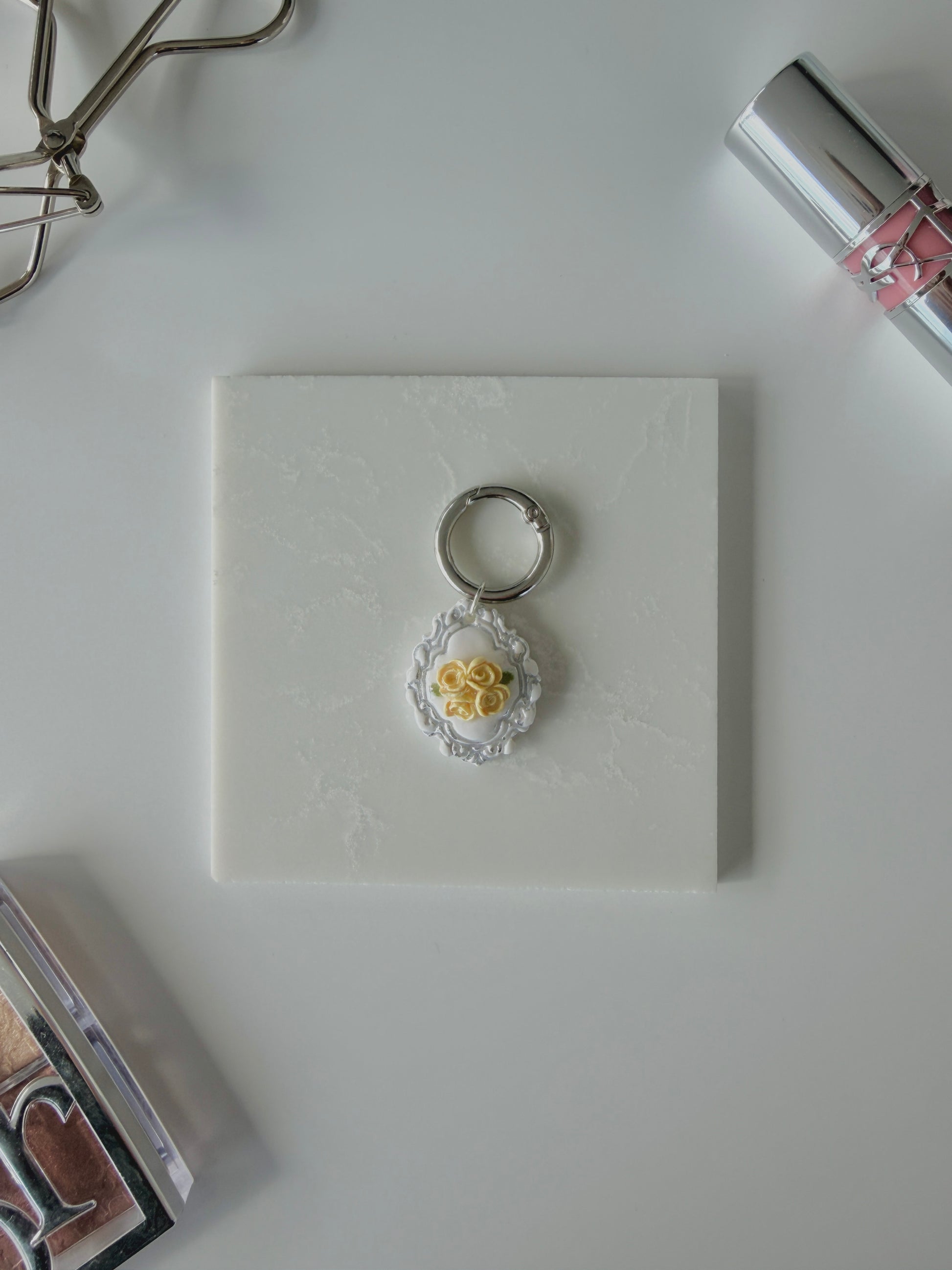 A product image of a white pet ID Tag with four yellow roses and silver engraving attached to a silver ring sits on a piece of marble. Staged around it are a silver eyelash curler, YSL lipstick, and Dior makeup palette all on a white background.