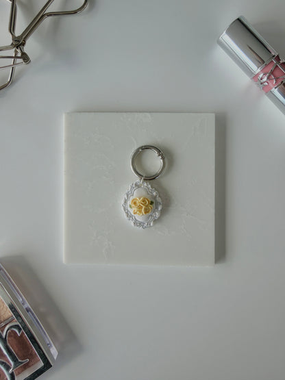 A product image of a white pet ID Tag with four yellow roses and silver engraving attached to a silver ring sits on a piece of marble. Staged around it are a silver eyelash curler, YSL lipstick, and Dior makeup palette all on a white background.