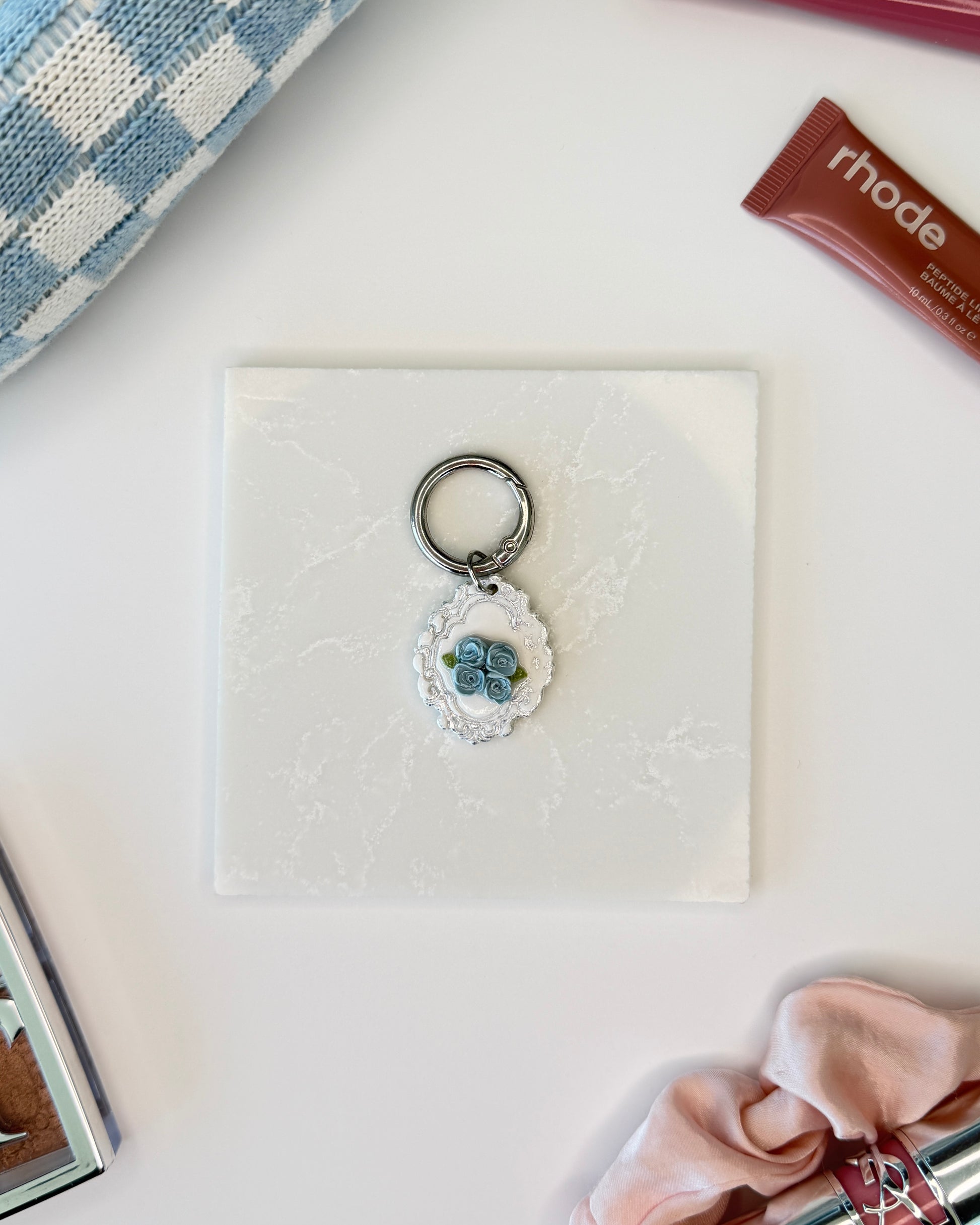 A product image of a white pet ID Tag with four blue roses and silver engraving attached to a silver ring sits on a piece of marble. Staged around it are a blue and white checkered makeup bag, pink satin scrunchie, rhode lip gloss, YSL lipstick, and Dior makeup palette all on a white background.