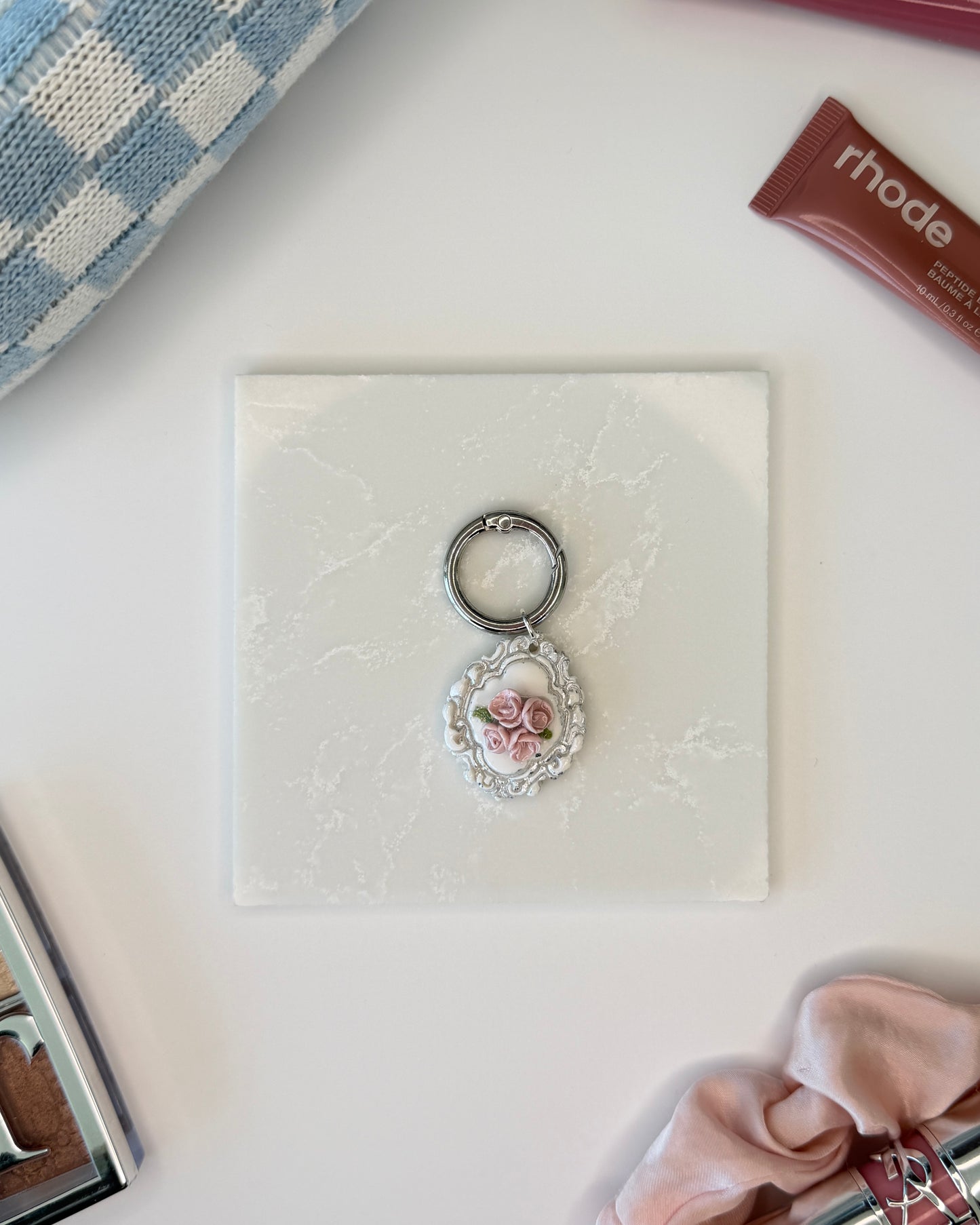 A product image of a white pet ID Tag with four pink roses and silver engraving attached to a silver ring sits on a piece of marble. Staged around it are a blue and white checkered makeup bag, pink satin scrunchie, rhode lip gloss, and Dior makeup palette all on a white background.