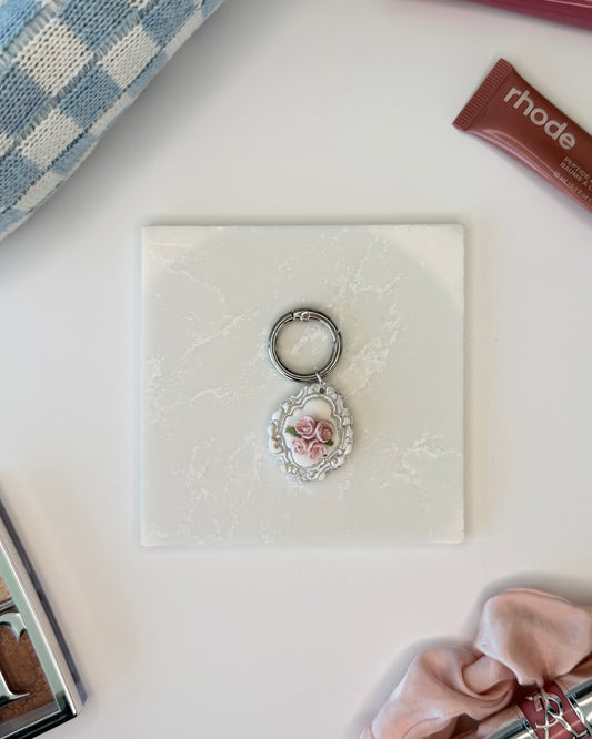 A product image of a white pet ID Tag with four pink roses and silver engraving attached to a silver ring sits on a piece of marble. Staged around it are a blue and white checkered makeup bag, pink satin scrunchie, rhode lip gloss, and Dior makeup palette all on a white background.