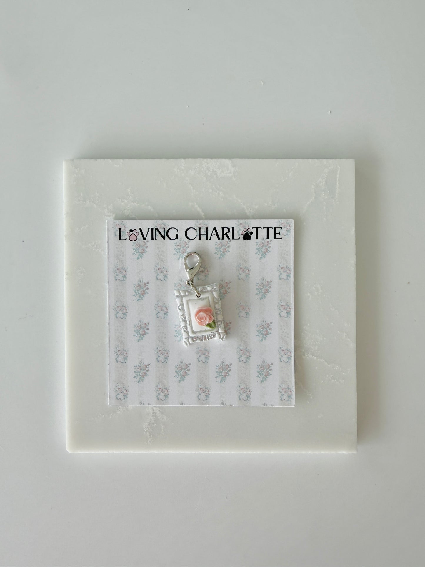 Small pet tag with silver engraving and single pink rose on a card with 'Loving Charlotte' branding on a light gray background