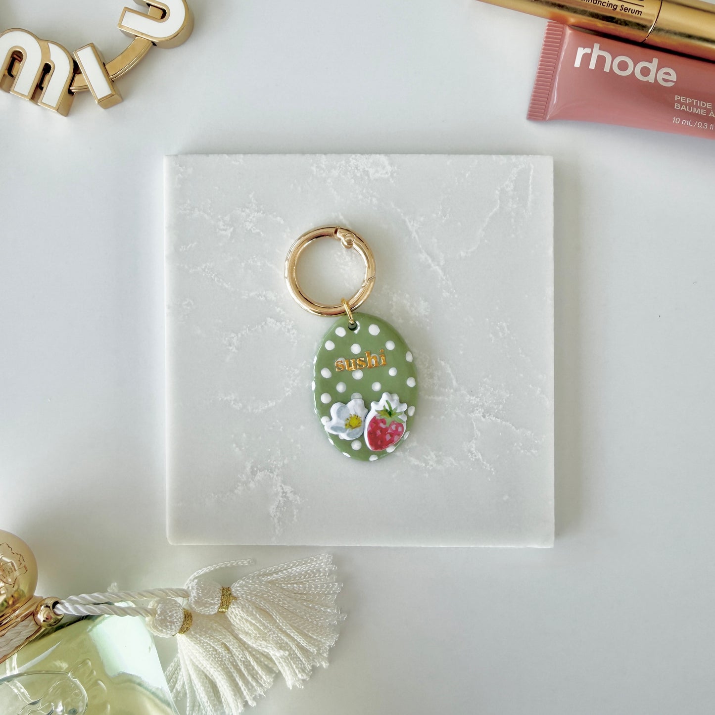 Pet tag with gold lettering ("sushi") on an oval strawberry design with white polka dots laying on a slab of marble next to cosmetics.