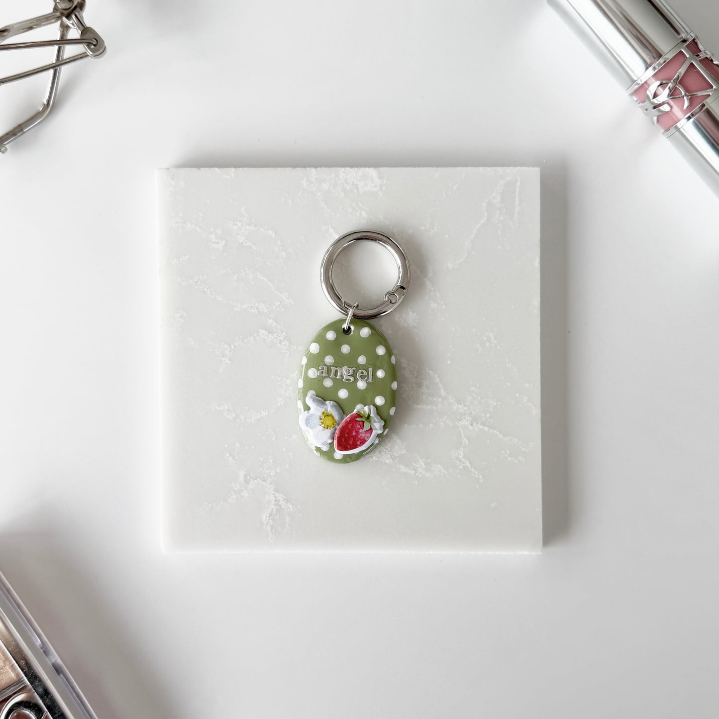 Pet tag with silver lettering ("angel") on an oval strawberry design with white polka dots laying on a slab of marble next to cosmetics.