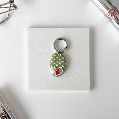 Pet tag with silver lettering ("angel") on an oval strawberry design with white polka dots laying on a slab of marble next to cosmetics.